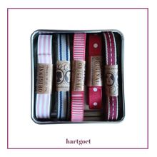 Load image into Gallery viewer, A square metal tin containing four decorative ribbon bands in pink, blue, red, and burgundy, each with a tan paper label reading ORIGINAL. The tin is centered on a white background with a thin purple border, and the word hartgoet appears below.