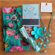 Load image into Gallery viewer, A flat-lay of a coordinated gift set on a wooden surface, including a turquoise floral scarf, a notebook with print reading 'Happiness Never Goes Out of Style,' a flower-shaped turquoise hair clip, a clear tube with manicure essentials and a turquoise beaded necklace.