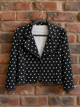Load image into Gallery viewer, A black cropped jacket with white polka dots hanging on a black hanger against a red-brown brick wall.