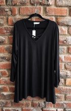 Load image into Gallery viewer, A black long-sleeve V-neck top hanging on a black hanger against a red-brown brick wall.
