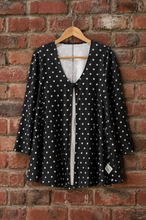 Load image into Gallery viewer, A black long-sleeve cardigan with white polka dots, hanging on a wooden hanger against a red brick wall.