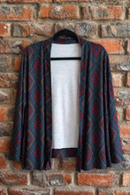 Load image into Gallery viewer, A long-sleeve open-front cardigan with a dark navy and red geometric diamond pattern, hanging on a black hanger against a rustic red brick wall.