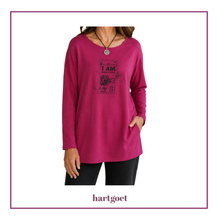 Load image into Gallery viewer, A woman wearing a long-sleeve magenta pink tunic top with a black graphic print on the front that reads 'I AM' surrounded by decorative elements. The image is framed with a purple border, and the brand name 'hartgoet' is printed at the bottom.