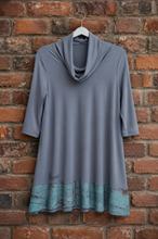Load image into Gallery viewer, A gray cowl-neck tunic top with three-quarter sleeves, featuring a distressed teal and brown patterned hem, hanging on a white hanger against a red brick wall.