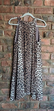 Load image into Gallery viewer, A sleeveless leopard print dress, hanging on a grey hanger against a red-brown brick wall.
