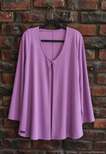 Load image into Gallery viewer, A lavender purple long-sleeve top with a split V-neckline, hanging on a black hanger against a red-brown brick wall.