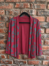 Load image into Gallery viewer, A maroon open-front cardigan with a teal floral pattern, hanging on a black hanger against a rustic red brick wall.