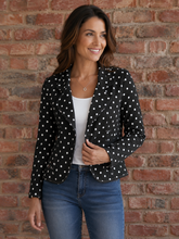 Load image into Gallery viewer, A woman wearing a black long-sleeve blazer with white polka dots, styled over a white top and blue jeans, standing against a red-brown brick wall.