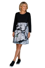 Load image into Gallery viewer, A woman modeling a long-sleeve knee-length dress with a solid black upper section and a black, white, and gray abstract print lower skirt, standing against a white background.