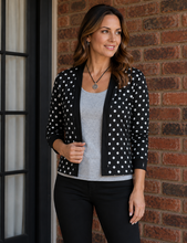 Load image into Gallery viewer, A woman wearing a black cardigan with white polka dots over a light gray tank top and black pants, standing in front of a red brick wall.
