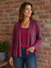 Load image into Gallery viewer, A woman wearing a burgundy open-front cardigan with a teal floral pattern over a solid burgundy top and blue jeans, standing against a red brick wall.
