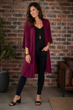 Load image into Gallery viewer, Model wearing a long maroon jacket, black top and pants, with a brickwall background