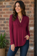 Load image into Gallery viewer, A woman wearing a long-sleeve burgundy top and dark blue jeans, standing in front of a brick wall with a potted plant on the left.