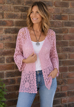 Load image into Gallery viewer, A woman wearing a light pink open-front crochet cardigan over a white tank top and blue jeans, standing against a red-brown brick wall.