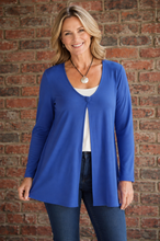 Load image into Gallery viewer, A smiling blonde woman wearing a purple long-sleeve open-front cardigan over a white top and dark blue jeans, standing against a red-brown brick wall.