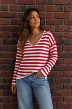 Load image into Gallery viewer, A woman wearing a red and white horizontally striped long-sleeve V-neck top with light blue jeans, standing against a red-brown brick wall.