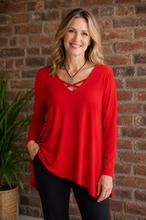 Load image into Gallery viewer, A woman wearing a bright red long-sleeve top with a criss-cross V-neckline detail, paired with black pants, standing in front of a rustic brick wall with a potted plant on the left.