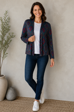 Load image into Gallery viewer, A woman modeling a navy blue cardigan with a red and teal geometric pattern, worn open over a white t-shirt and dark blue jeans, standing in a styled indoor setting with a textured beige wall, potted plants, and a woven rug.