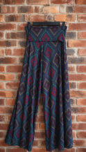 Load image into Gallery viewer, A pair of wide-leg pants with a colorful geometric diamond pattern, hanging on a black hanger against a red brick wall.
