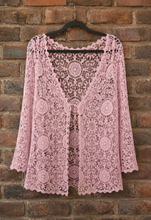 Load image into Gallery viewer, A pink open-front cardigan with long sleeves and circular crochet patterns, hanging on a black hanger against a rustic red-brown brick wall.