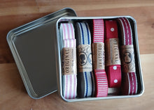 Load image into Gallery viewer, A metal tin containing five decorative ribbon straps in different color and pattern combinations, with kraft paper bands labeled 'ORIGINAL' around each ribbon, placed on a wooden surface.