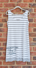 Load image into Gallery viewer, A sleeveless white dress with gray horizontal stripes, featuring the text 'A GIRL MUST BE 2 THINGS Classy & Fabulous' in gray script and print near the bottom left, finished with a gray lace hem. The dress is hanging on a grey hanger against a red-brown brick wall.