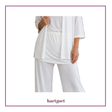 Load image into Gallery viewer, A model wearing a white two-piece lounge set consisting of a short-sleeve top, hip skirt and matching wide-leg pants, framed by a purple border with the brand name hartgoet at the bottom.