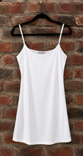 Load image into Gallery viewer, A white spaghetti-strap camisole top hanging on a black hanger against a red-brown brick wall.