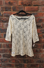 Load image into Gallery viewer, A light yellowy lace top with 3/4 sleeves hanging on a black hanger against a red brick wall background.