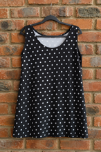 Load image into Gallery viewer, A sleeveless black dress with a white polka dot pattern, hanging on a black hanger against a red brick wall.
