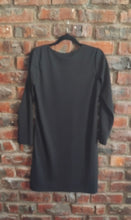 Load image into Gallery viewer, A black long-sleeve dress hanging on a black hanger against a red-brown brick wall.
