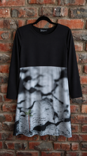 Load image into Gallery viewer, A black long-sleeve dress with a black and white cracked marble print lower half, hanging on a black hanger against a red brick wall.
