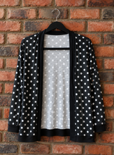 Load image into Gallery viewer, A black open-front cardigan with white polka dots hanging on a black hanger against a red-brown brick wall.
