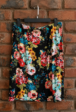 Load image into Gallery viewer, A black skirt with a vibrant multicolor floral print featuring red, pink, blue, yellow, and white flowers, hanging on a black hanger against a red brick wall.
