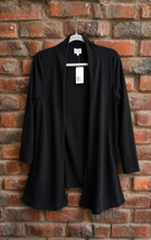 Load image into Gallery viewer, A black open-front cardigan with long sleeves, hanging on a hanger against a red-brown brick wall.

