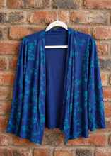 Load image into Gallery viewer, A blue open-front cardigan with a teal floral pattern, hanging on a white hanger against a red-brown brick wall.
