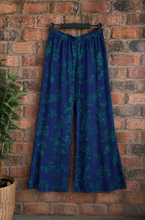 Load image into Gallery viewer, Blue wide-leg trousers with a teal floral pattern, hanging from a black hanger against a red brick wall.
