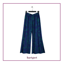 Load image into Gallery viewer, A pair of wide-leg pants in blue with a teal floral pattern, hanging on a black hanger against a white background with a purple border and the brand name hartgoet below
