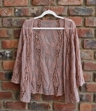 Load image into Gallery viewer, A brown lace cardigan hanging on a white hanger against a rustic red brick wall.
