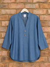 Load image into Gallery viewer, A blue chambray long-sleeve top with a split V-neck and three-quarter buttons, hanging on a white wooden hanger against a rustic red-brown brick wall.
