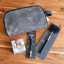 Load image into Gallery viewer, A gray textured zippered pouch lies on a wooden tabletop alongside several accessories: a small packaged set of two bottles and a glass container, a black-and-silver pen on a branded card, and a black-and-silver massage tool on a dark card.

