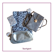 Load image into Gallery viewer, A flat-lay product photo of a small notebook, leopard print scarf, a gray faux-leather crossbody bag, and a silver gray make-ip bag, all on a white background with a purple border.
