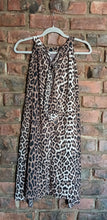 Load image into Gallery viewer, A sleeveless leopard print dress with a tie-neck detail at the back, hanging on a white plastic hanger against a red-brown brick wall.
