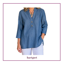 Load image into Gallery viewer, A woman wearing a blue chambray long-sleeve button-up top with white pants, shown from the neck down against a white background with a purple border. The brand name 'hartgoet' is printed at the bottom.
