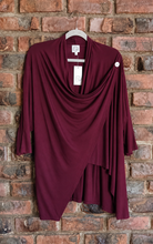 Load image into Gallery viewer, A draped burgundy long-sleeve top hanging on a white hanger against a rustic red-brown brick wall. The top features a cowl neckline, asymmetrical draped layers, and a small white button detail on the upper right shoulder.
