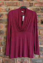 Load image into Gallery viewer, A long-sleeve burgundy top with a draped cowl neckline and twisted front detail, hanging on a black hanger against a rustic red-brown brick wall.
