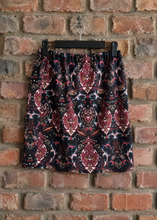 Load image into Gallery viewer, A black skirt with a red, blue, and white ornate paisley pattern, hanging on a black hanger against a red-brown brick wall.
