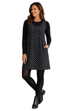 Load image into Gallery viewer, A woman modeling a black sleeveless shift dress with white polka dots over a black long-sleeve top, paired with black tights and black ankle boots, on a solid white background.
