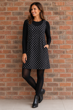 Load image into Gallery viewer, A woman models a black sleeveless mini dress with white polka dots over a black long-sleeve top, paired with black tights and black ankle boots, standing against a red-brown brick wall.

