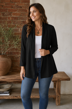 Load image into Gallery viewer, A woman wearing a black open-front long-sleeve cardigan over a white tank top and blue jeans, standing in a rustic indoor setting with a brick wall, wooden bench, and potted plant.
